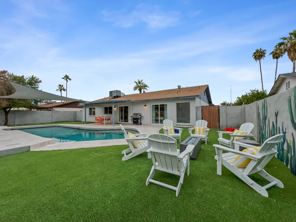 A cheerful backyard scene with a sparkling swimming pool, adirondack chairs, an outdoor dining area, and a decorative wall adorned with cacti. The space is illuminated by natural light, suggesting an inviting setting for both day and evening gatherings.