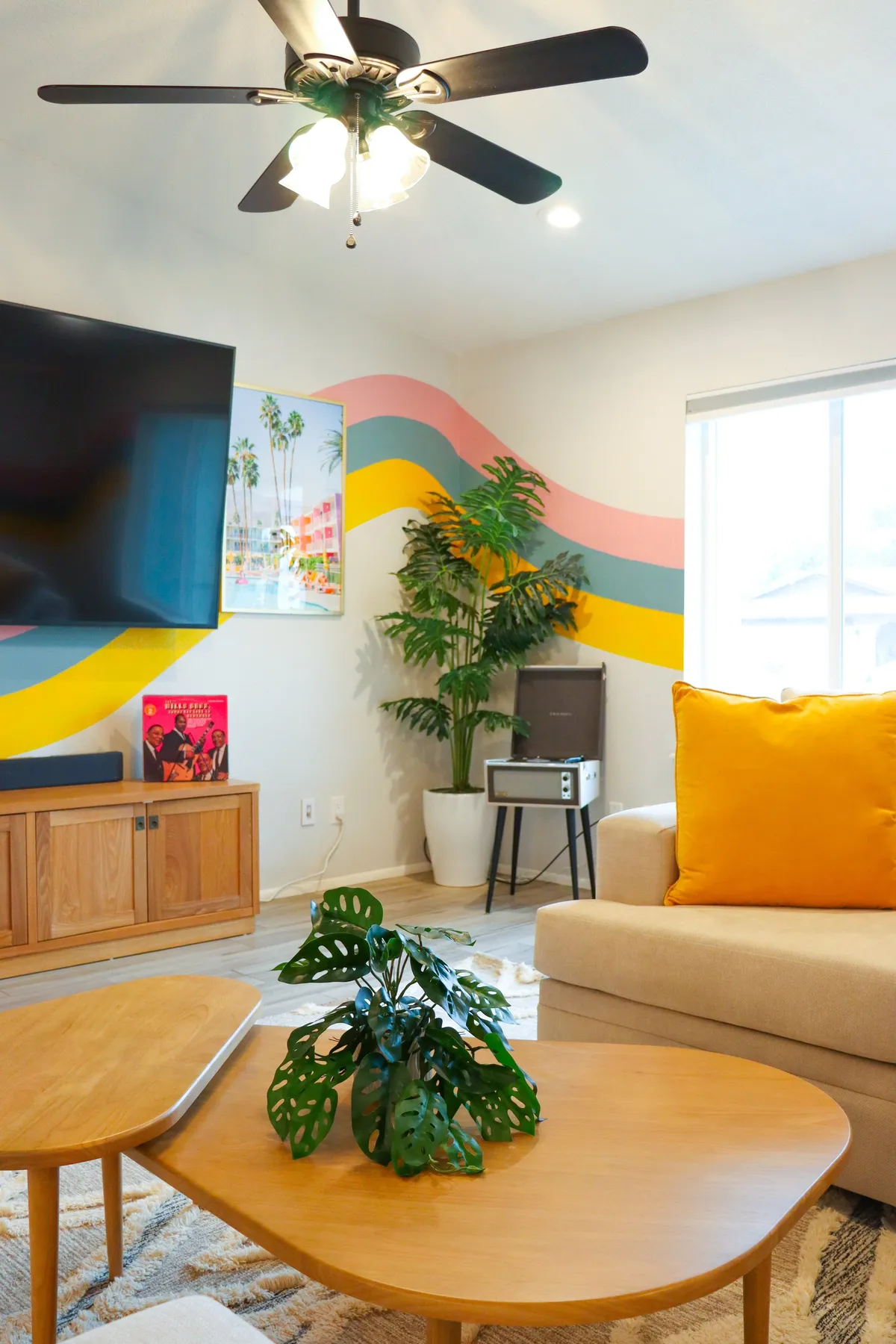 A spacious living room featuring a large flat-screen TV, colorful wall mural, stylish coffee table, and vibrant yellow accent pillow. The arrangement encourages gatherings and relaxation, with natural light streaming in through the windows and views extending towards the outdoors.