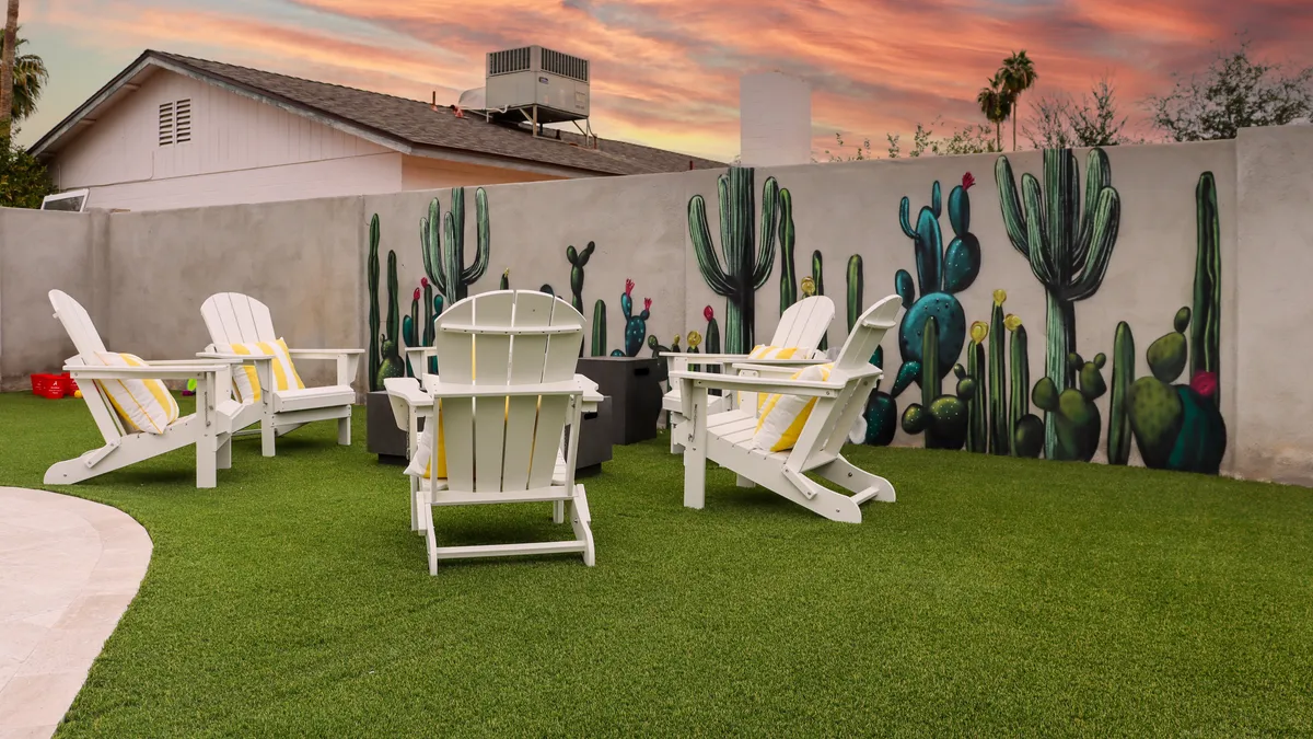 A cozy backyard scene featuring white Adirondack chairs arranged near a fire pit, artificial grass underfoot, a striking cactus mural, and a relaxed outdoor atmosphere ideal for socializing or unwinding. The space is designed for outdoor enjoyment and leisure, with elements that suggest a low-maintenance, recreational yard.