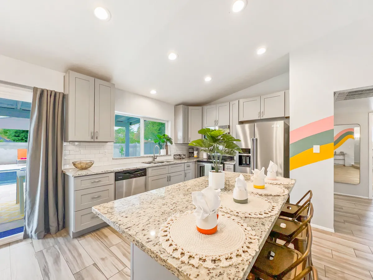 A cheerful kitchen scene with a granite-topped island, stainless steel appliances, colorful wall accents, and indoor plants. The space feels lively and modern, with ample room for both meal prep and socializing.