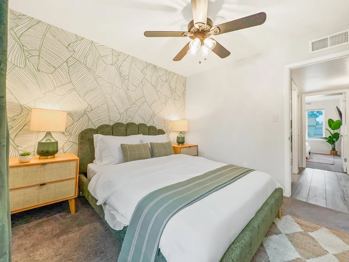 A cozy bedroom with a green accent wall adorned with a leaf pattern, a plush green headboard, and nightstands with glowing lamps. The bed is made up invitingly, evoking comfort and restful sleep. Soft, diffused daylight enhances the serene mood.