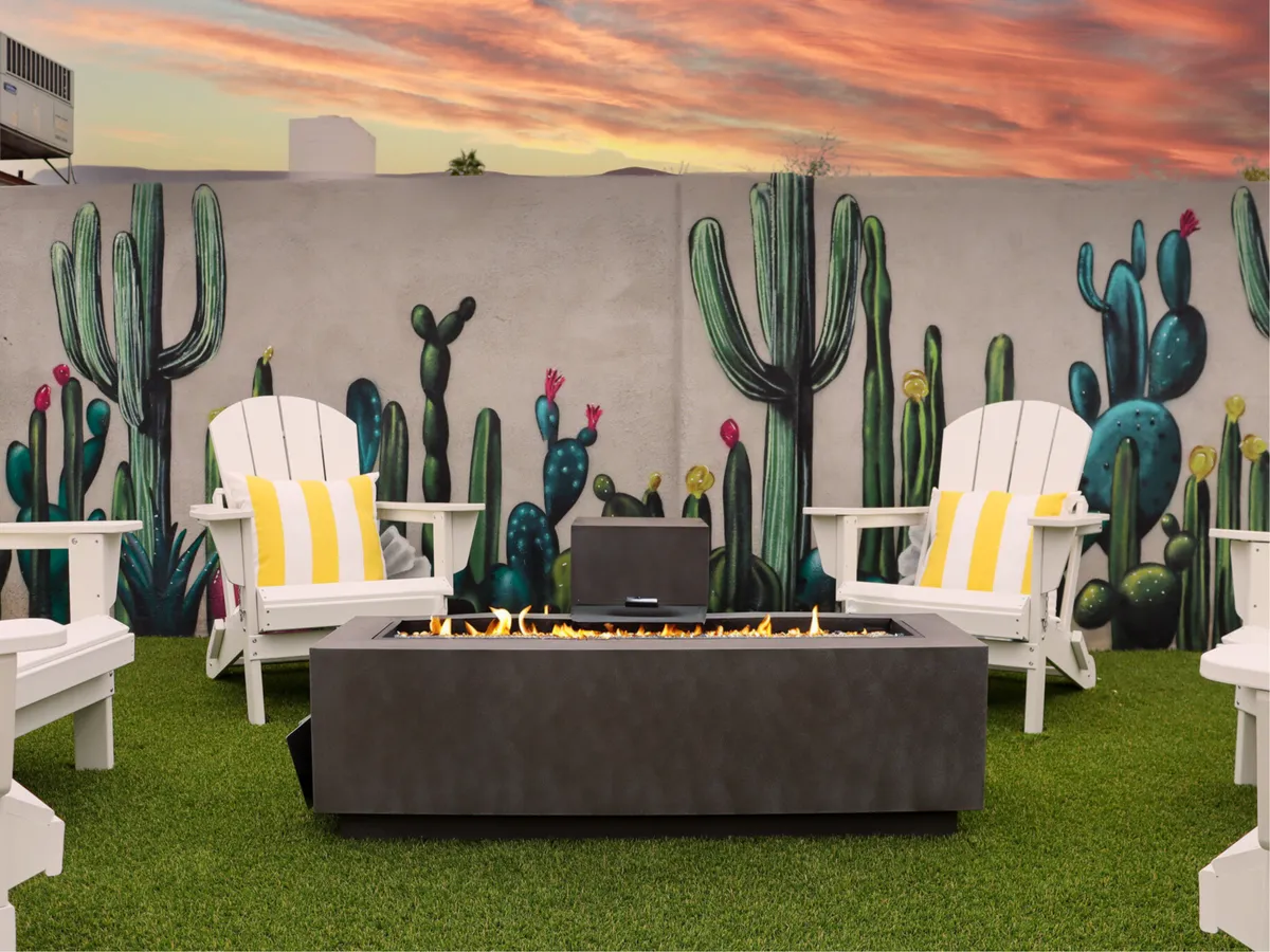 This cozy exterior image centers on a fire pit surrounded by Adirondack chairs, with a vibrant cactus mural as a backdrop. The inviting setup evokes an ideal spot for evening gatherings, storytelling, or marshmallow toasting as the sun sets.