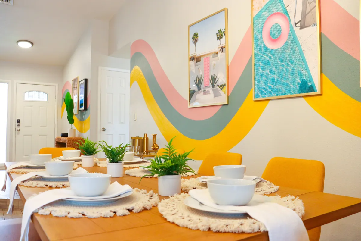 A brightly lit dining area features a table set for a meal, vivid wall art, and colorful décor. The lively mood suggests celebrations, family gatherings, and moments of togetherness, with a subtle connection to the outdoors through abundant natural light.