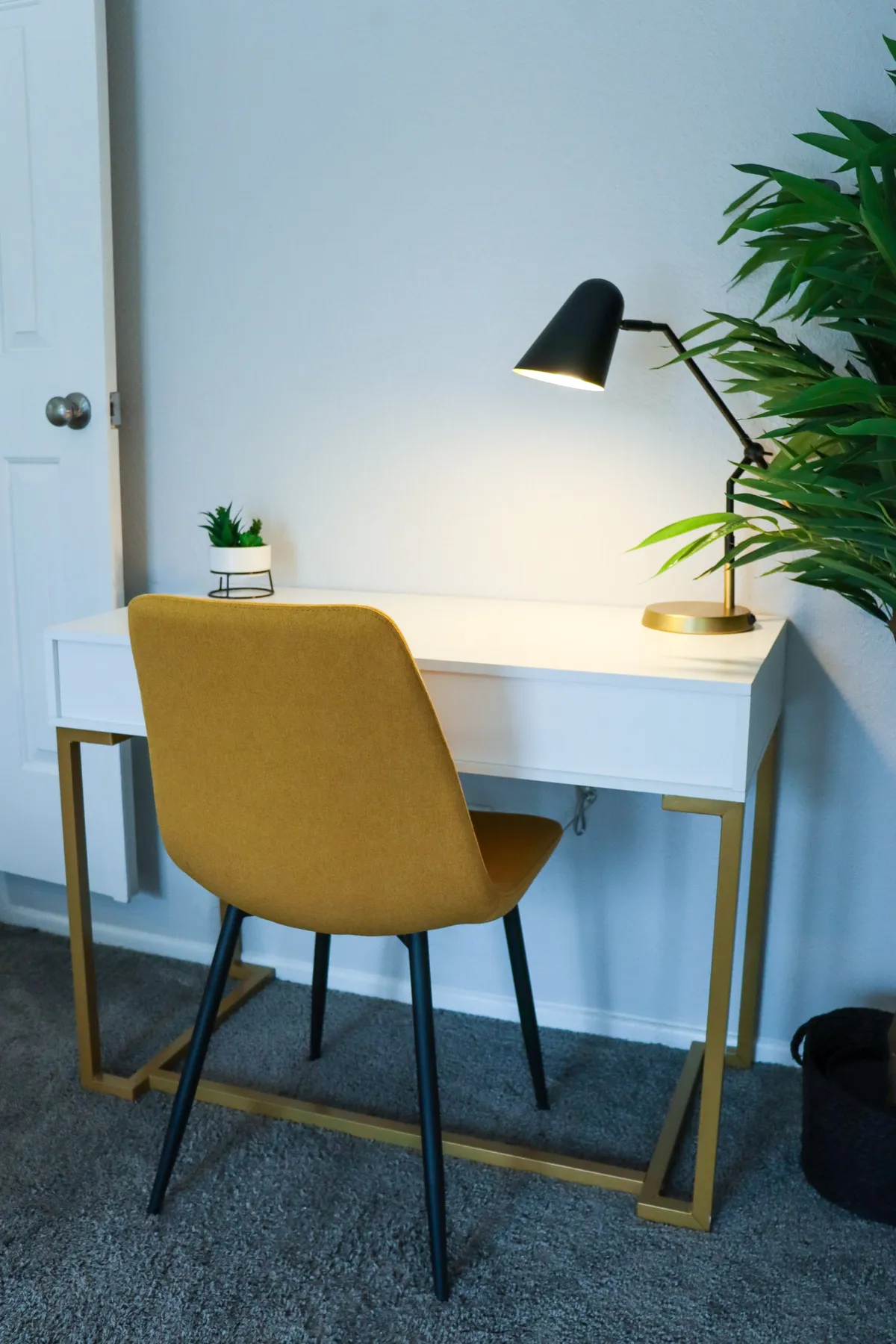 A modern, compact workspace with a comfortable desk and chair, lit by a stylish lamp. A leafy plant adds a touch of freshness, and natural light fills the corner, making it a peaceful spot for work or study.