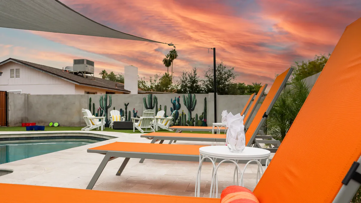 A vibrant exterior scene featuring a sparkling swimming pool surrounded by sleek orange lounge chairs. In the background, a playful cactus mural decorates the wall, while the sky glows with warm sunset hues. The space is designed for both relaxation and lively poolside gatherings.
