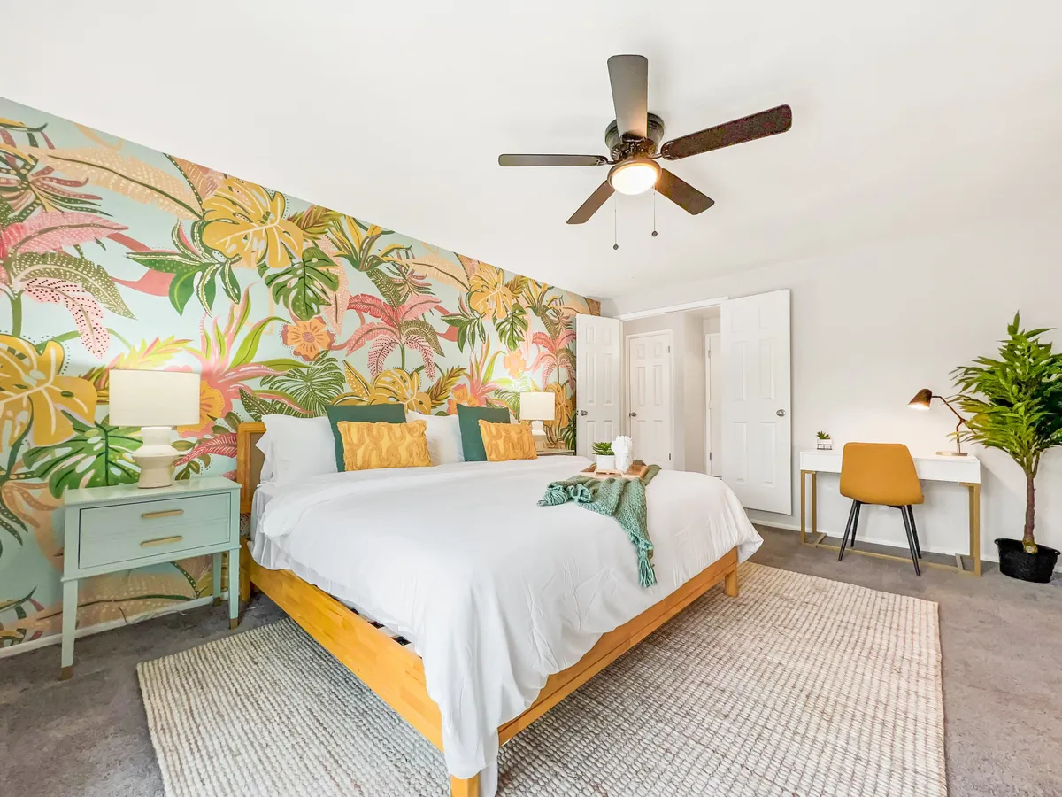 A tranquil master bedroom featuring a king-size bed with crisp linens, tropical wallpaper, nightstands with soft lighting, a writing desk perfect for remote work, and a lush green plant by the window. Natural light streams in, creating a welcoming and serene retreat.