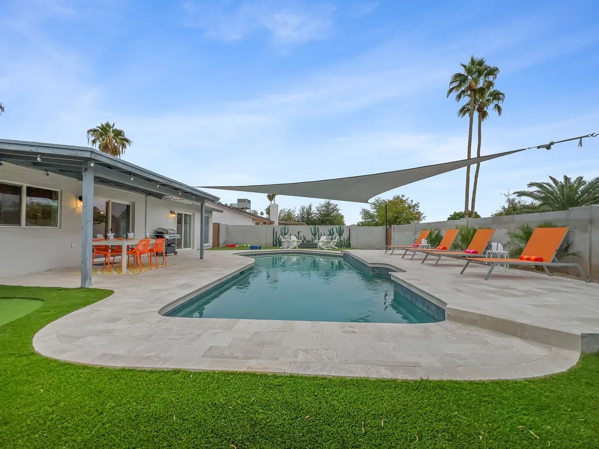 An expansive outdoor view showcasing a swimming pool, shaded by a sail, with several lounge chairs and an adjacent outdoor dining area. The setup suggests a flexible space for swimming, sunbathing, dining, or socializing, all in a private backyard setting.