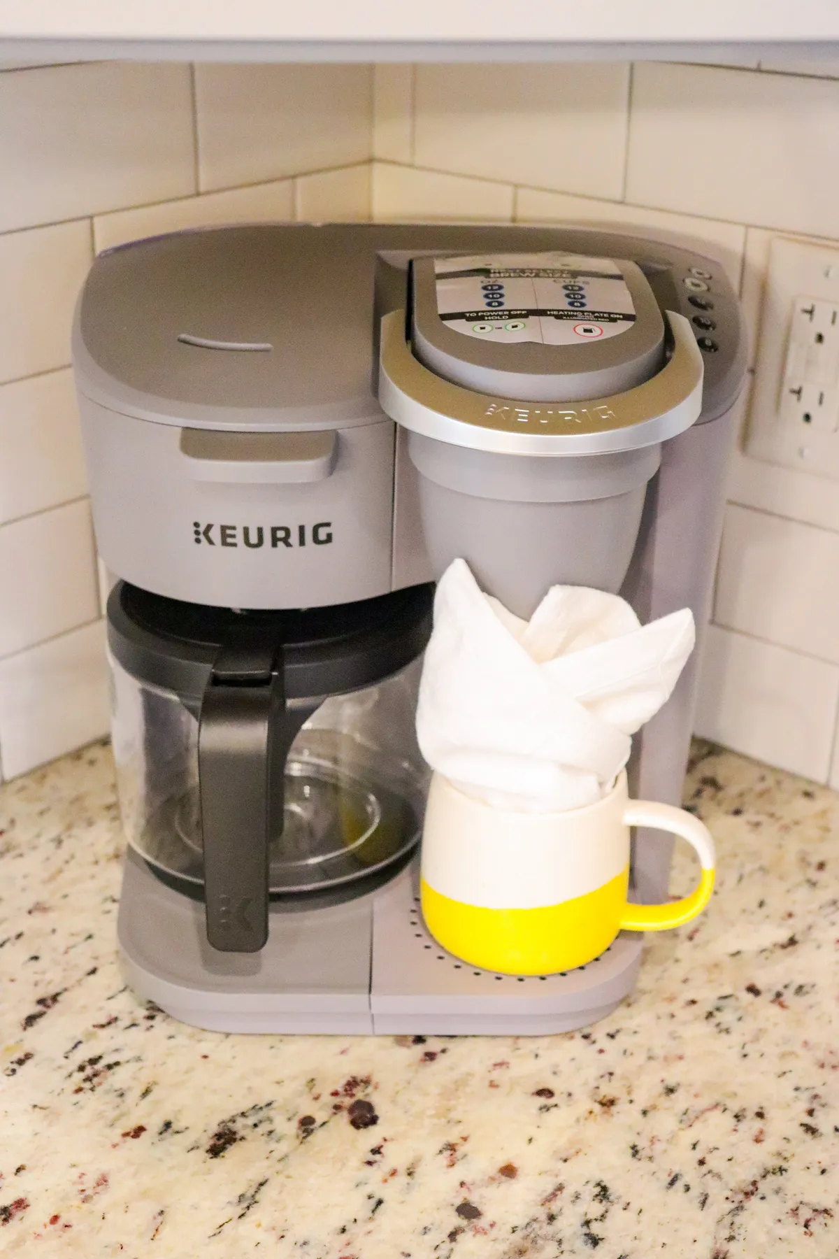 A close-up shot of a Keurig coffee maker on a kitchen counter, accompanied by a cheery yellow-accented mug and a glass carafe. The coffee setup sits against a bright, inviting backdrop, suggesting a convenient spot to start your day or take a break during work-from-home sessions.