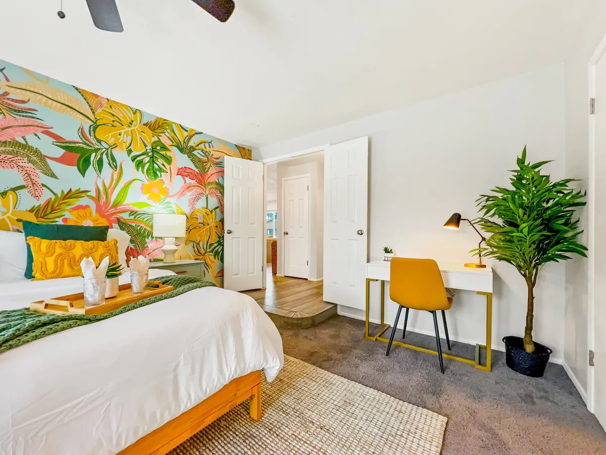 A bright bedroom with colorful floral wallpaper, a comfortable bed adorned with decorative pillows, and a minimalist desk setup. The workspace is cozy yet functional, ideal for remote work, reading, or relaxing in a cheerful environment.