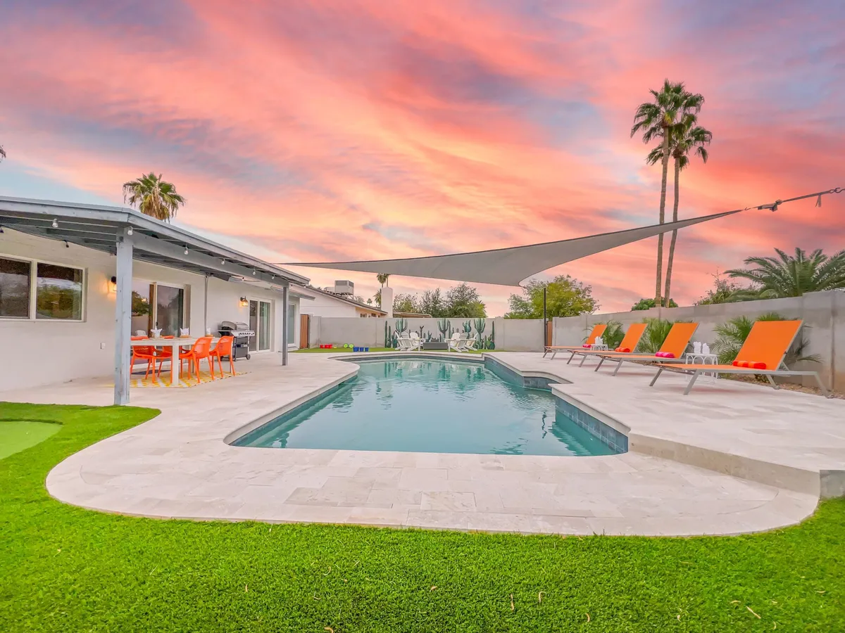 This vibrant exterior image showcases a sparkling swimming pool surrounded by lounge chairs, with an outdoor dining area visible in the background and a colorful sunset sky overhead. The scene captures the essence of relaxed poolside living, inviting guests to imagine themselves swimming, sunbathing, or enjoying an outdoor meal.