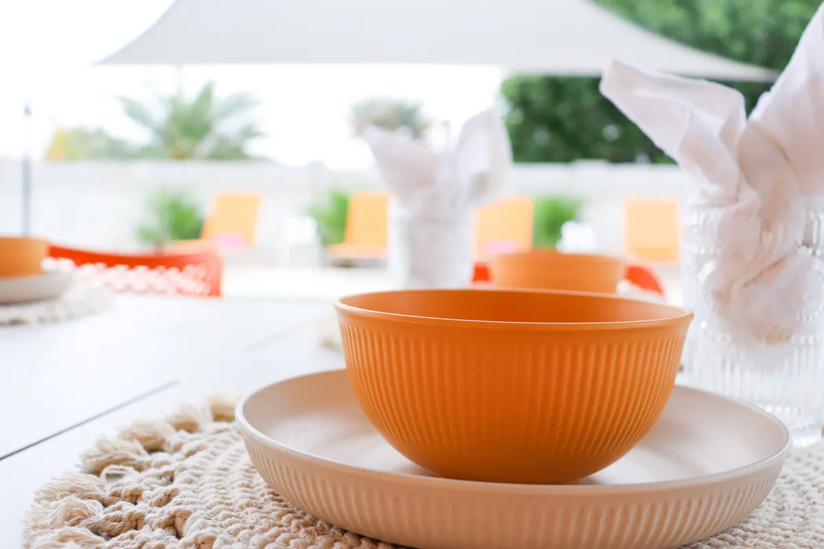A dining table set with a vibrant orange bowl and woven placemat sits in the foreground, while large windows offer a clear view onto an inviting outdoor space just beyond. The scene is bathed in natural light, suggesting easy movement between indoor and outdoor living.