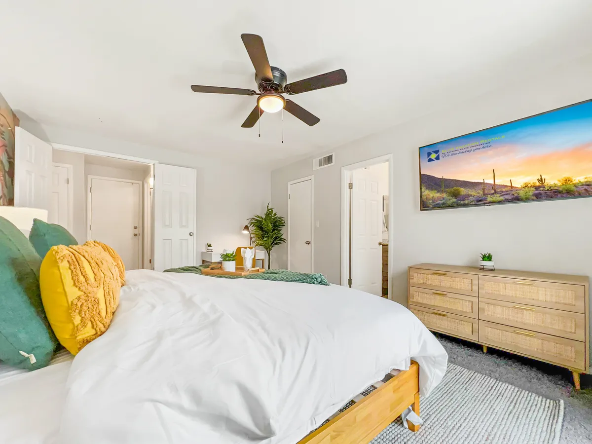 A comfortable bedroom with a bed dressed in decorative pillows, a ceiling fan above, a wall-mounted smart TV, dresser, and a leafy plant. Sunlight filters through the window, creating a calm, inviting space for relaxation and entertainment.