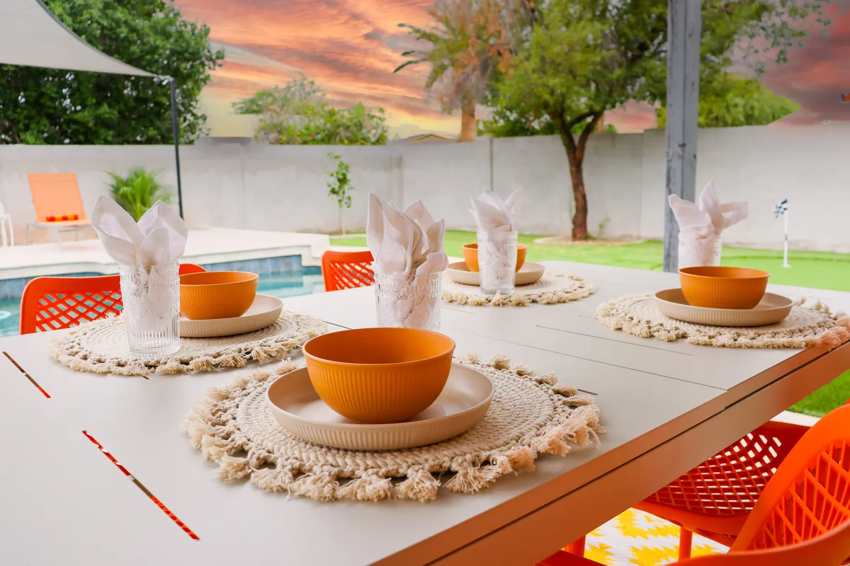 This image captures a cheerful outdoor dining space overlooking a swimming pool under a sunset sky. The table is set for a friendly gathering, evoking festive meals, celebrations, and shared stories in an inviting backyard setting.