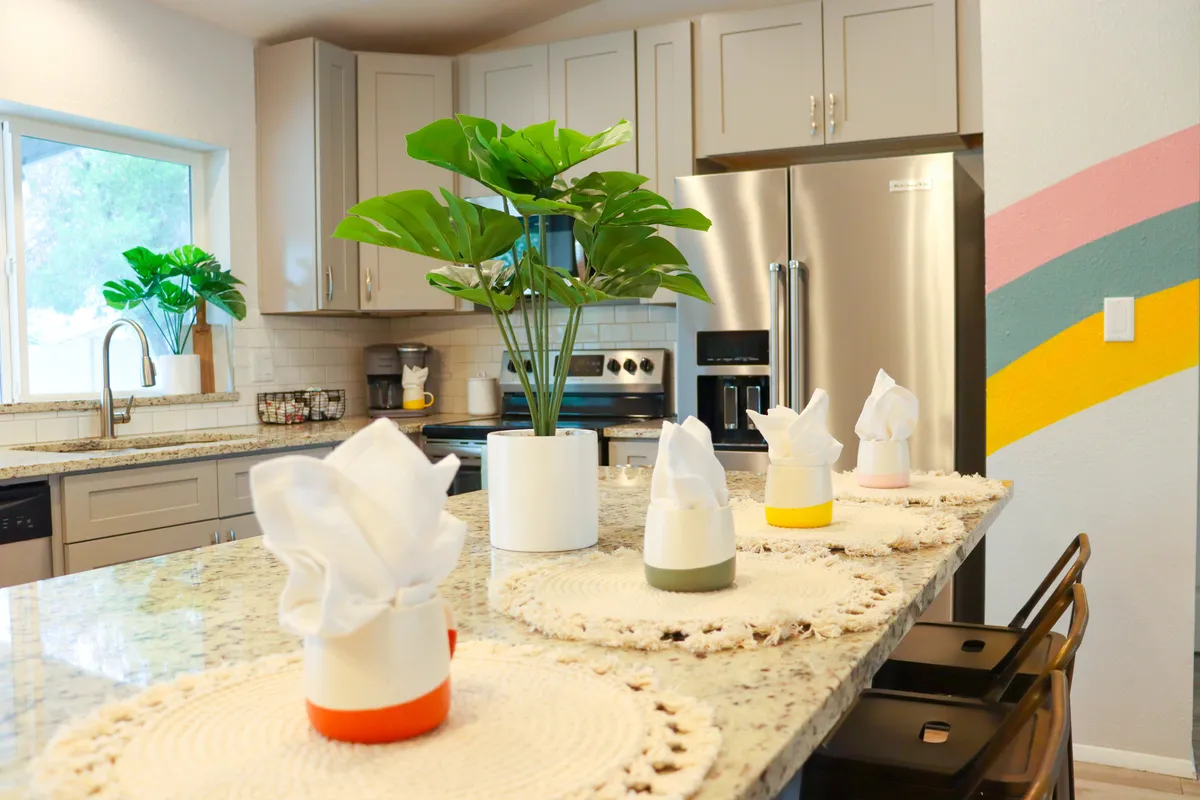 A spacious kitchen featuring a granite island, colorful napkin holders, and modern appliances. The atmosphere is lively yet relaxed, with hints of greenery adding freshness and life to the space. The layout suggests a communal area ideal for preparing meals together or hosting casual gatherings.