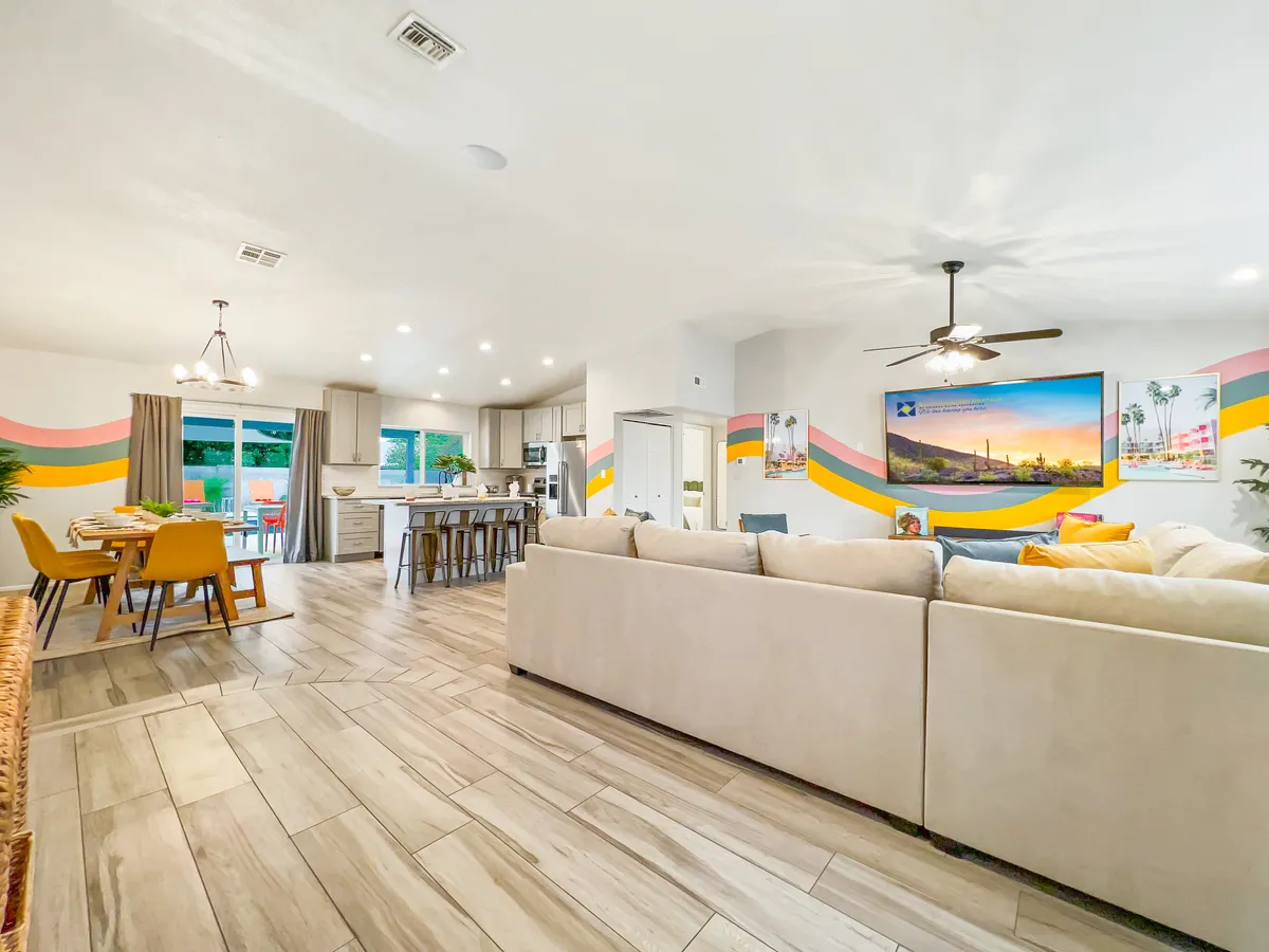 A family-friendly living space featuring a large sectional sofa, dining table with colorful chairs, wall-mounted TV with scenic artwork, and decorative wall painting. The space appears ready for family gatherings, casual meals, and entertainment, exuding a warm, welcoming mood.