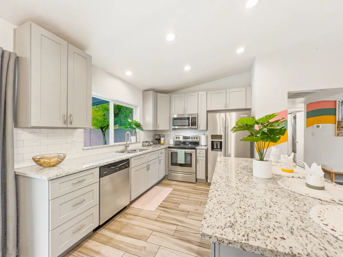This image showcases a modern kitchen with sleek granite countertops, stainless steel appliances, a window offering lush greenery views, and a decorative plant on the counter. The space is bright, inviting, and clearly designed for both cooking and entertaining guests.